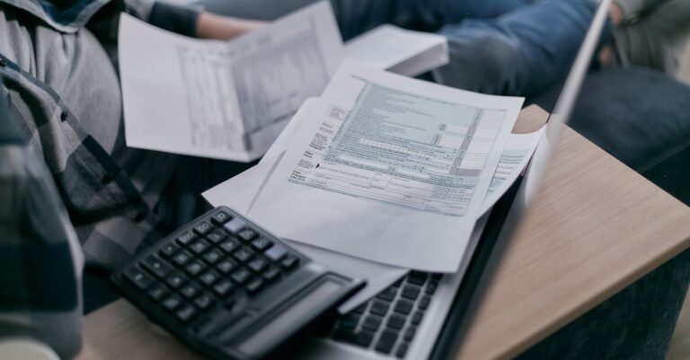 A young adult using a calculator and laptop to manage finances at home, surrounded by papers.