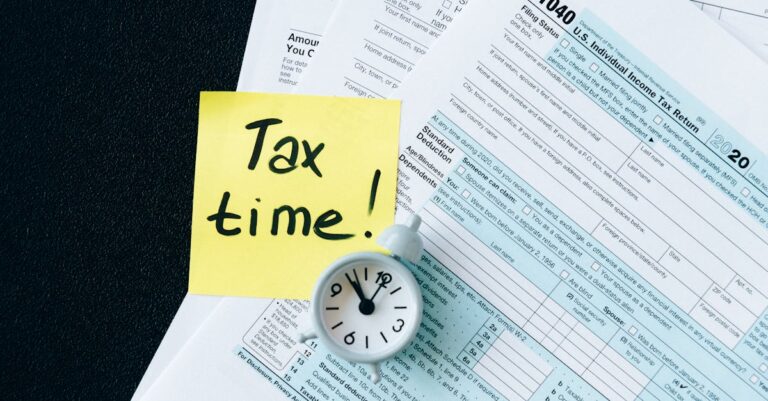 A stack of tax forms with a clock and yellow sticky note saying 'Tax time!' indicating urgency.