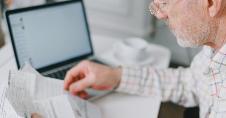 Elderly man wearing eyeglasses reading documents next to a laptop at home.