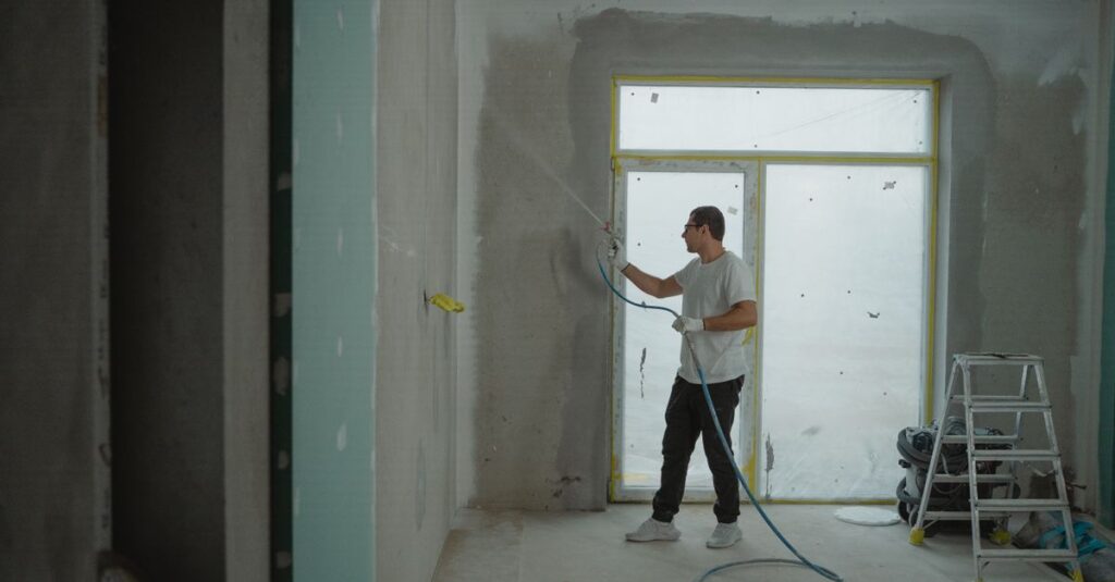 A man sprays paint on a wall while renovating an indoor room with professional equipment.