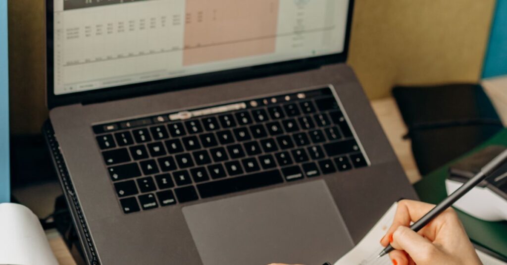 Hands writing notes beside a laptop displaying data in a bright office setup.