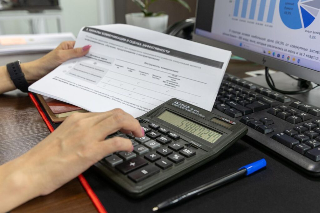 A person calculates financial data using a calculator and document, working at an office desk.